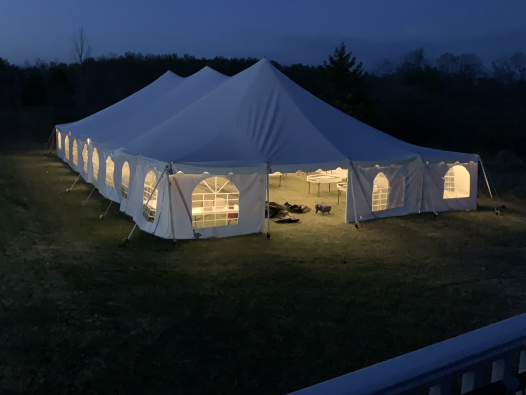 A large white canvas tent in the evening, lit from the inside, setup underway.